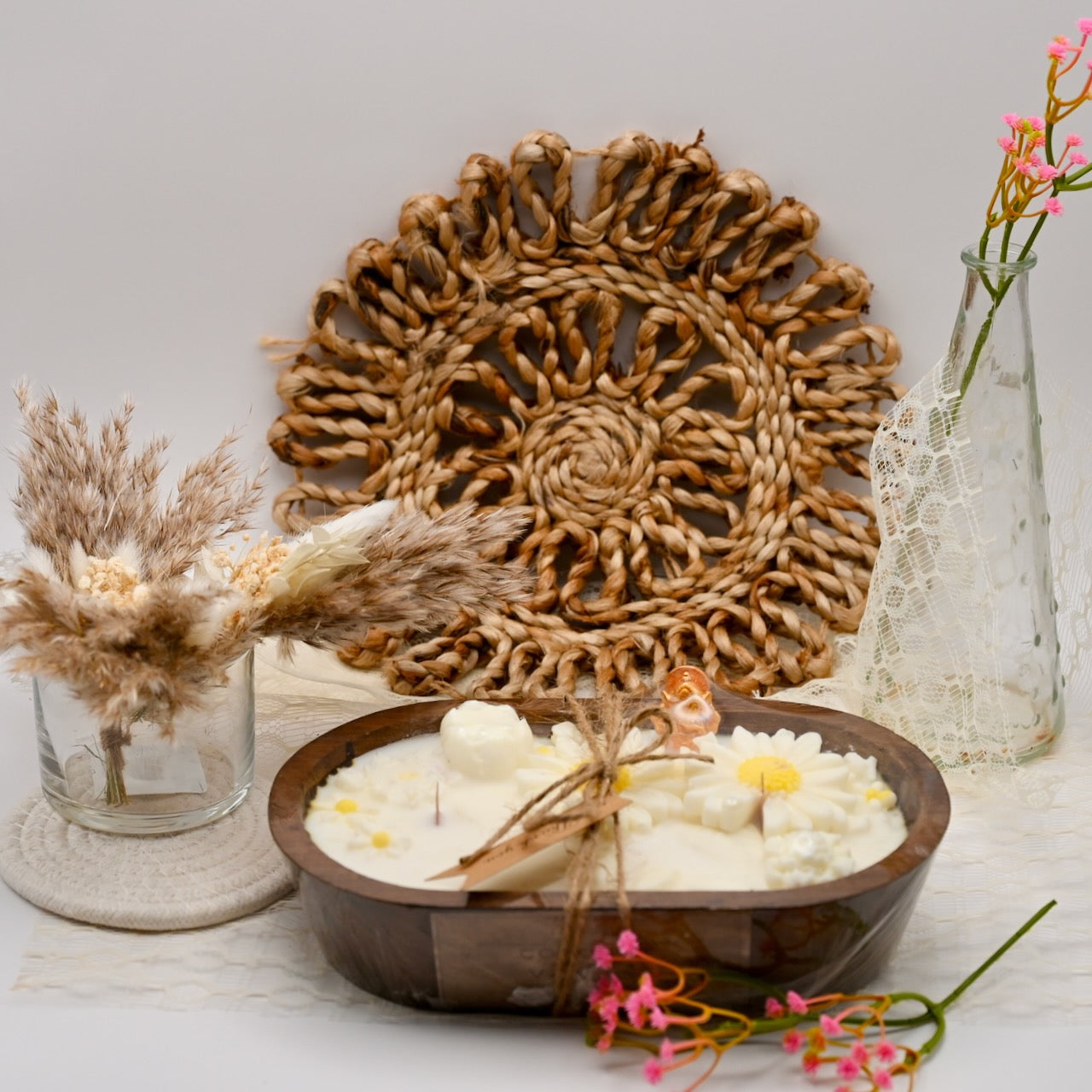Decorative setting with a woven wall art piece, candles in a wooden bowl, and floral elements on a light background.