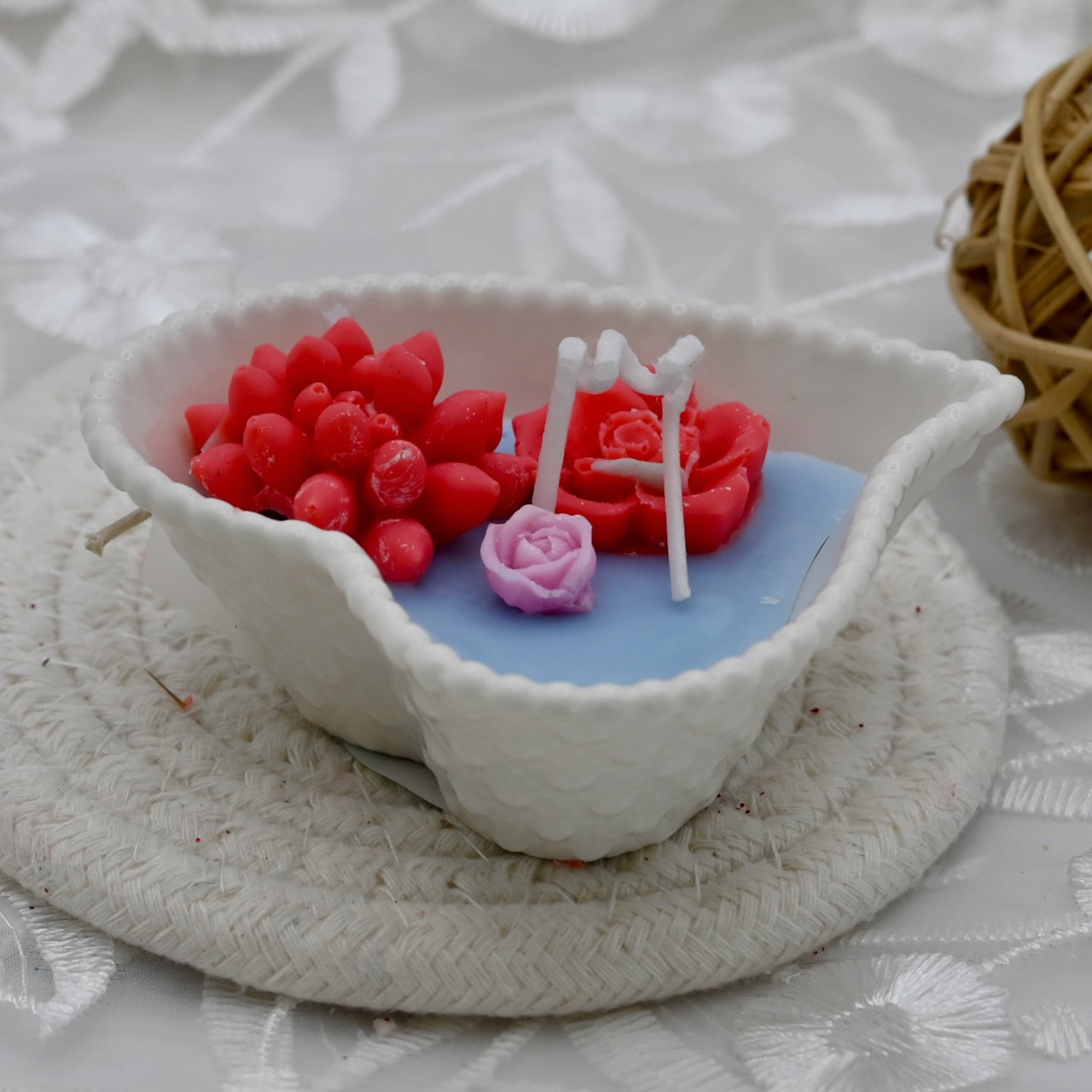 Decorative candle with red flowers and a pink rose in a white ceramic bowl on a textured surface.