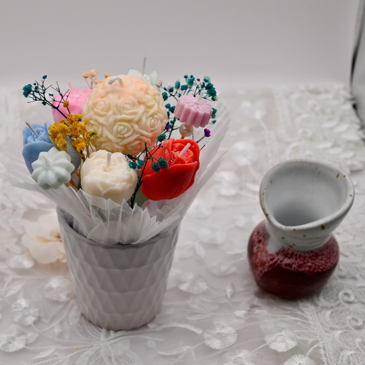 Bouquet of colorful soap flowers in a white container with a small red vase on a textured surface.