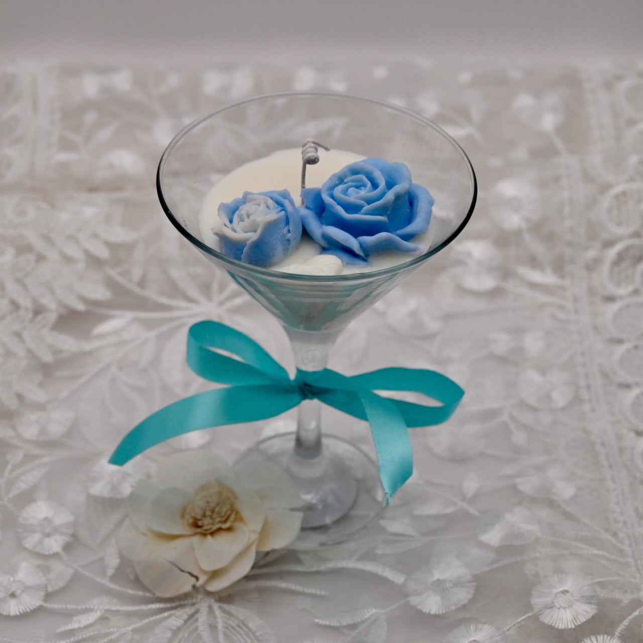 Decorative candle in a glass holder with blue roses and a turquoise ribbon on a floral-patterned background.