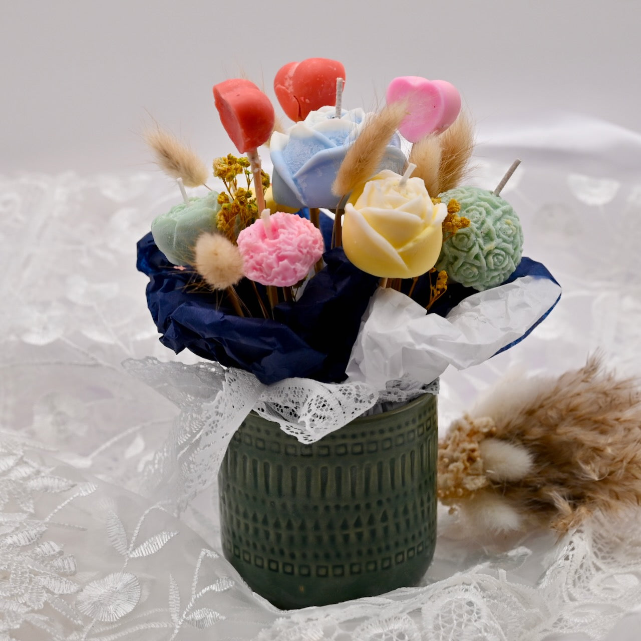 Decorative arrangement of colorful candles shaped like flowers in a green vase on a white lace background.