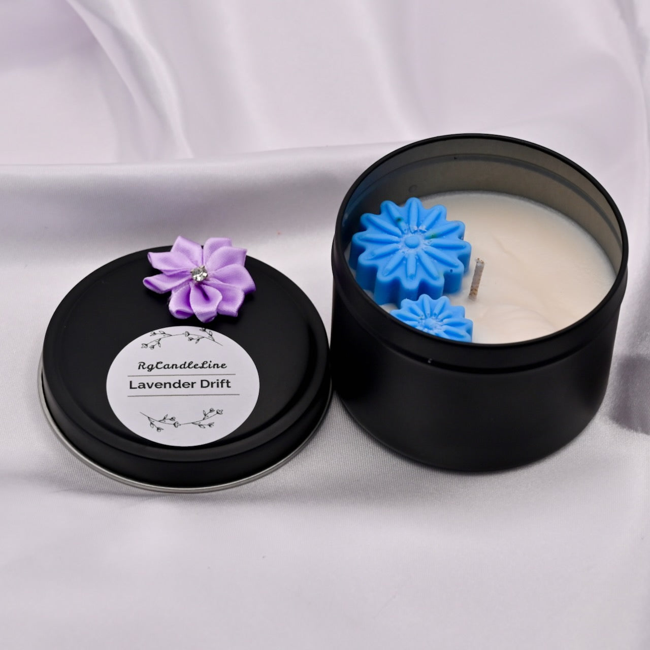 Black candle tin with a candle inside, featuring a purple flower-shaped wick and a blue flower-shaped candle insert on a white background.