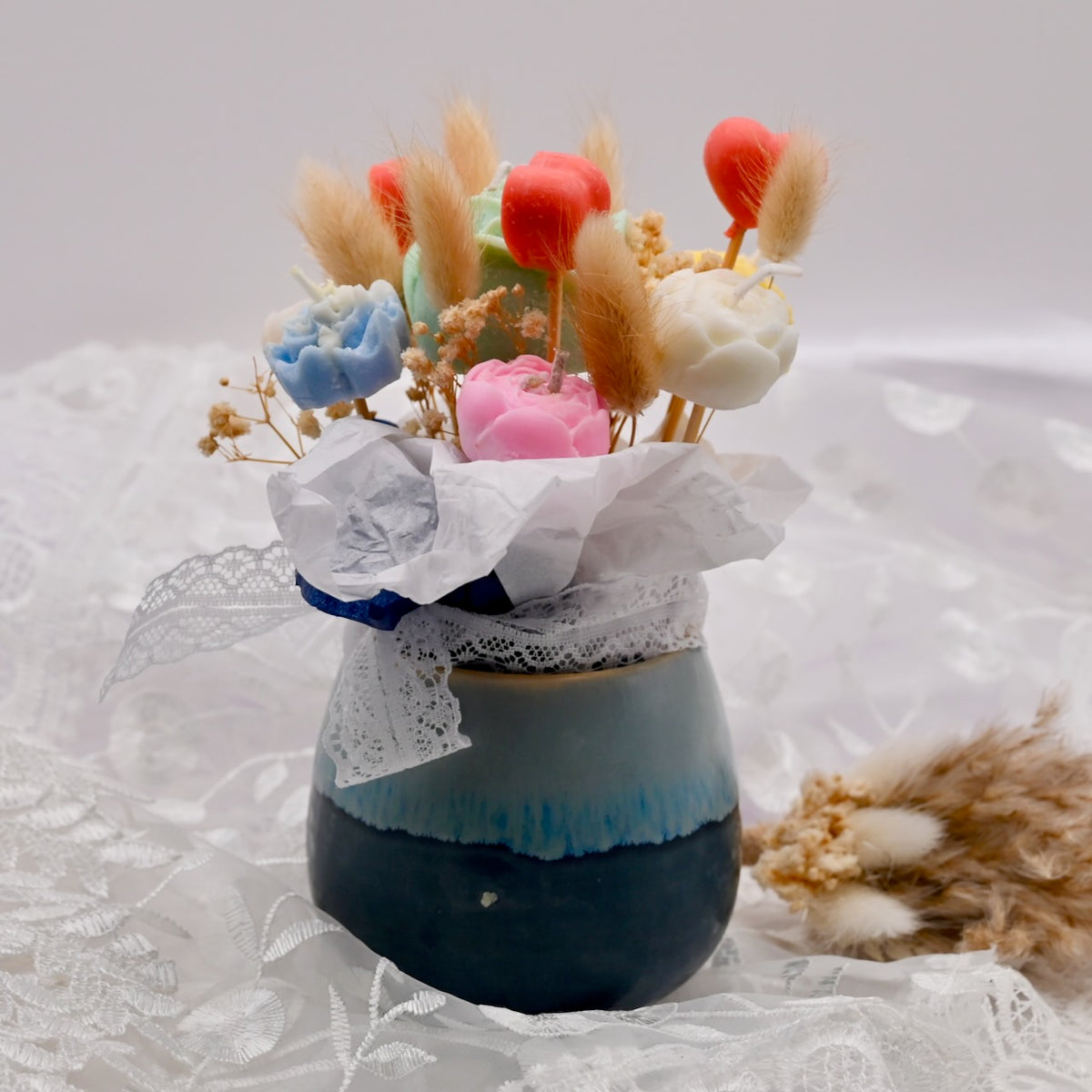 Vase arrangement of colorful soy wax flower candles with a lace wrap on a white lace background