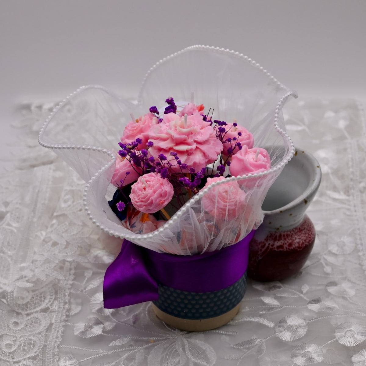 Soy wax flower candle arrangement in a pot with a purple ribbon on a lace tablecloth