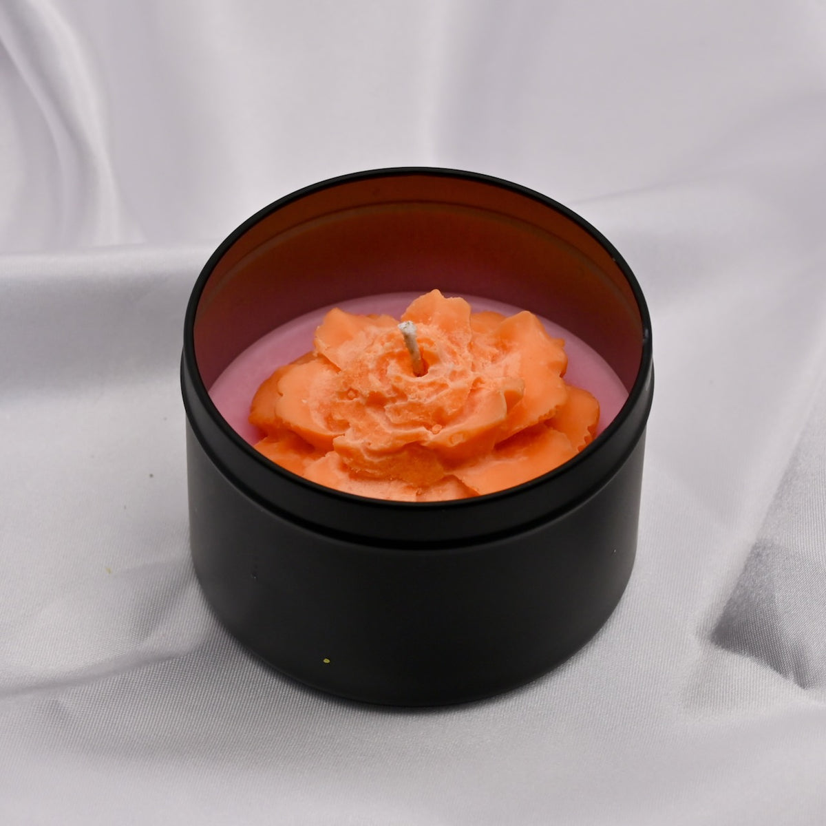 Black container with a pink candle and orange rose-shaped candle inside on a white background