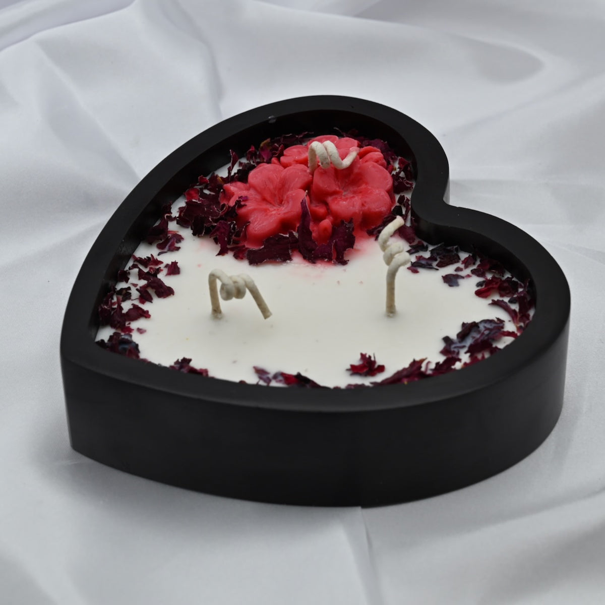 Angled view of a Blushing Heart soy wax candle in a black heart-shaped wooden container with red wax flowers and dried rose petals.