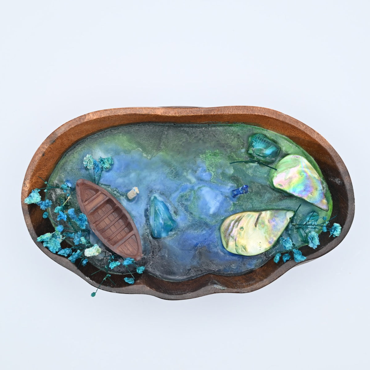Overhead view of the Whispering Sandal ocean bloom soy wax candle in a handcrafted wooden tray with blue-green water wax, a boat, shell accents, and dried blue flowers.