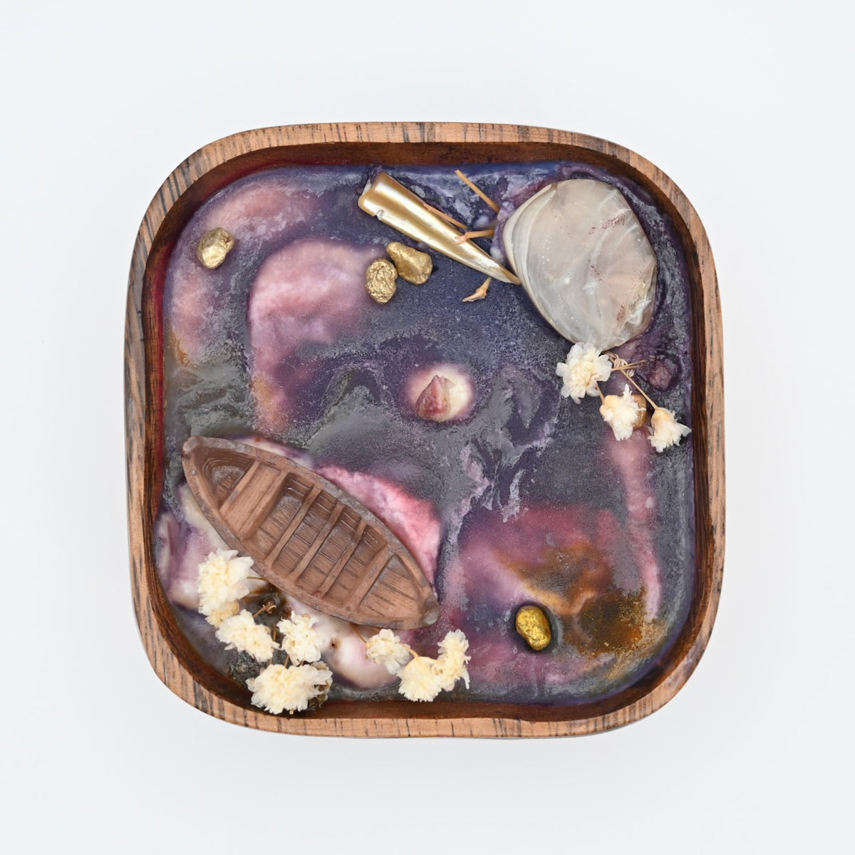 Top view of wooden bowl candle with purple wax, shell accents, dried flowers, and boat detail